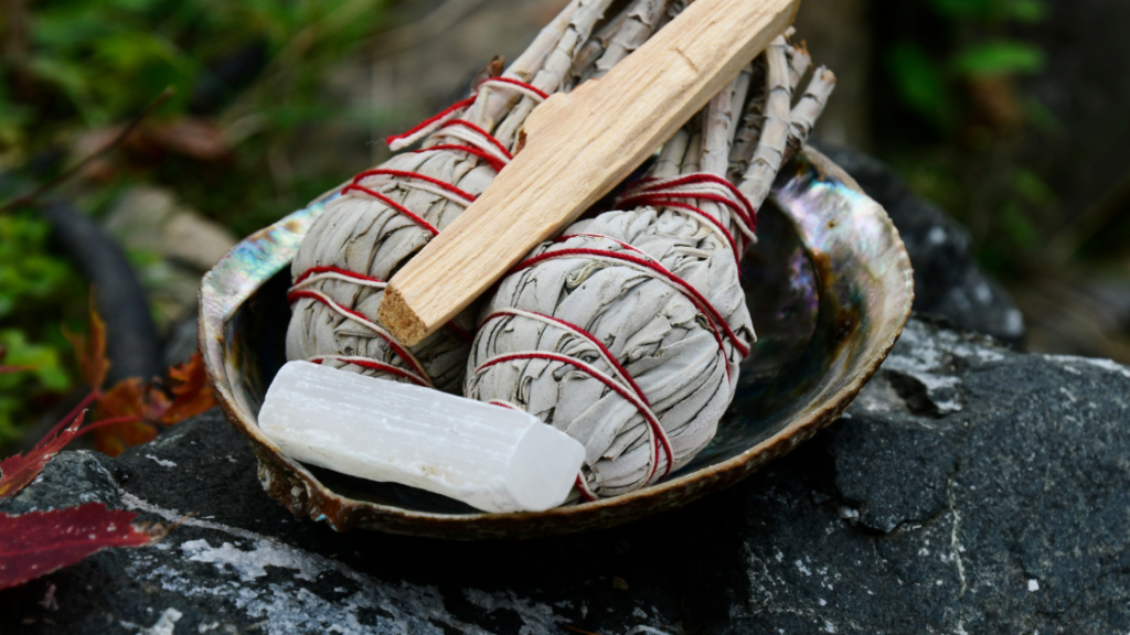 White sage bundles with red string and a selenite wand in an abalone shell on a rocky surface.